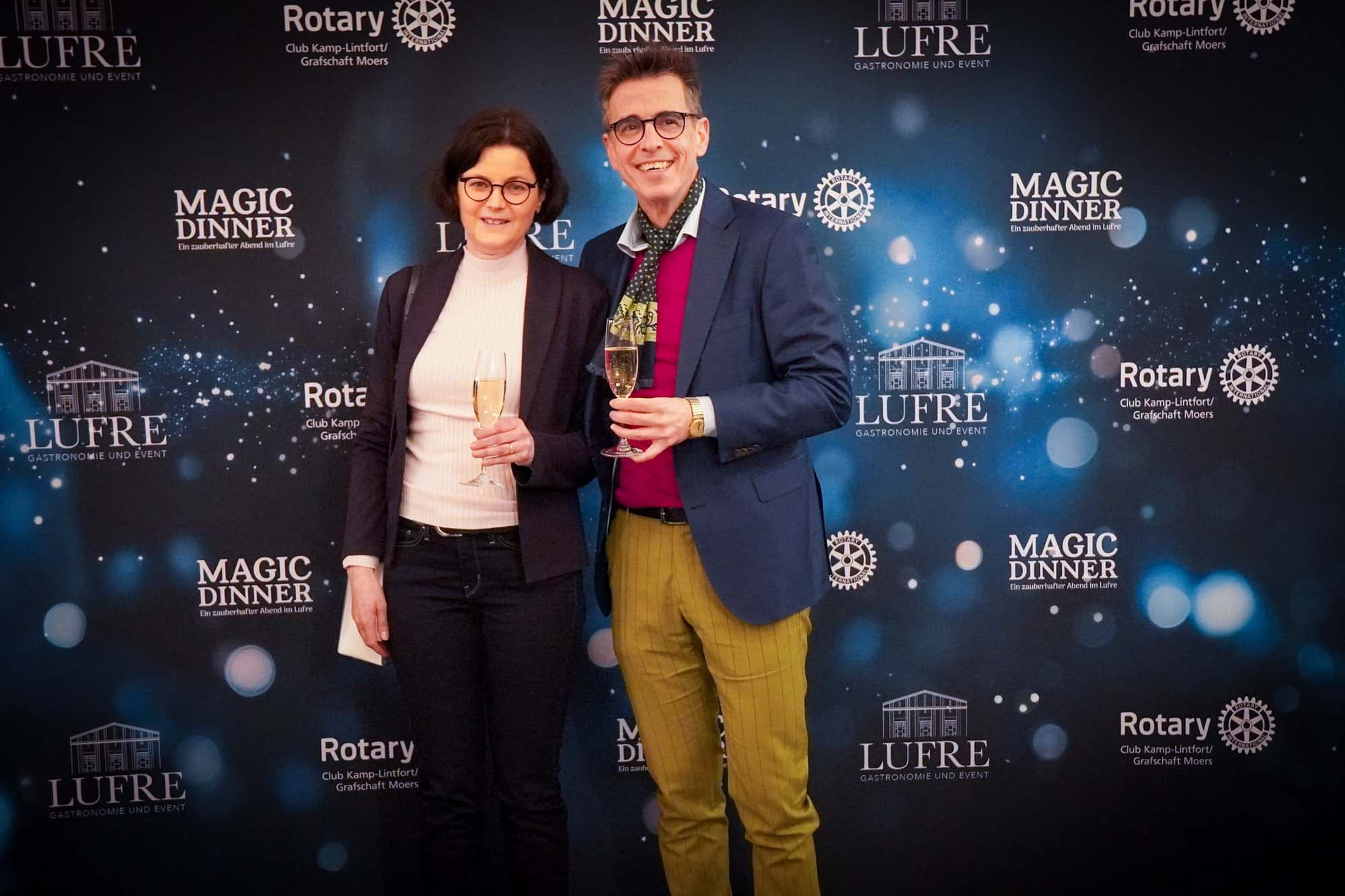 Ein elegant gekleidetes Paar mit Sektgläsern vor der Pressewand, sie in dunklem Blazer, er mit Halstuch und senfgelber Hose.